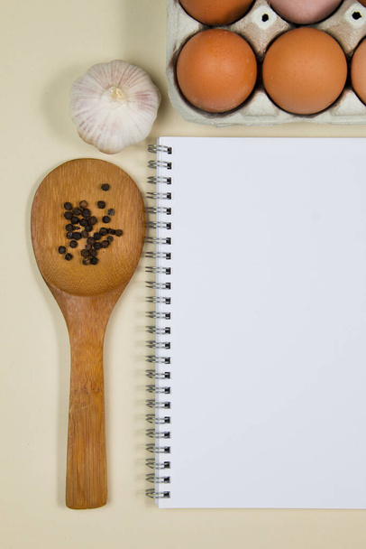 Houten pollepels met specerijen, eieren en knoflook met een blanco notebookpagina - Foto, afbeelding