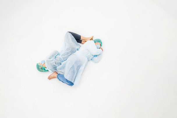 Zwei Frauen in medizinischen Schutzanzügen und Gesichtsmasken. Junge Mädchen liegen in medizinischen Schutzmänteln, Mützen und Masken auf dem weißen Boden. Vollständiger Schutz vor dem Pandemie-Virus - Foto, Bild