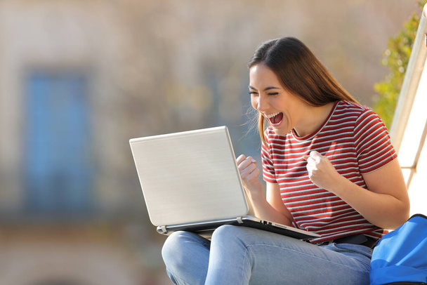 Excited student checking good news on laptop sitting outdoors in a university campus - Photo, image