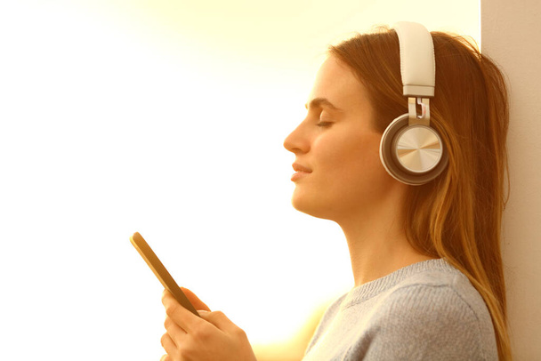 Side view portrait of a relaxed girl with closed eyed listening to music wearing headphones and holding smart phone at sunset - Foto, imagen