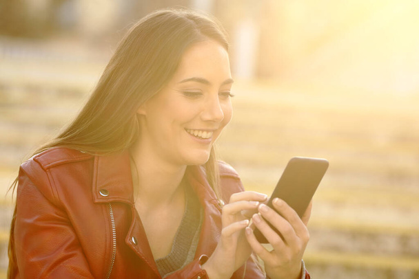 Onnellinen nainen käyttää älypuhelinta istuu kadulla lämpimässä valossa
 - Valokuva, kuva