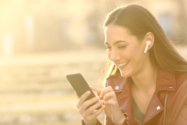 Happy woman wearing earbuds listening to music searching content on mobile phone with a warm light - Фото, изображение
