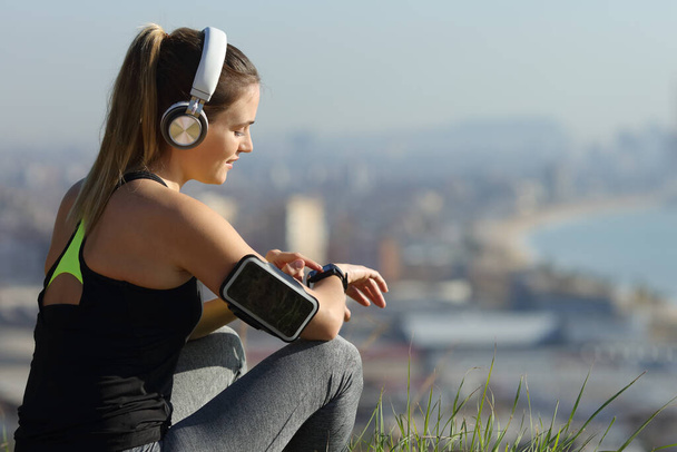 Runner wearing headphones checks music on smartwatch outdoors in city outskirts - Фото, зображення