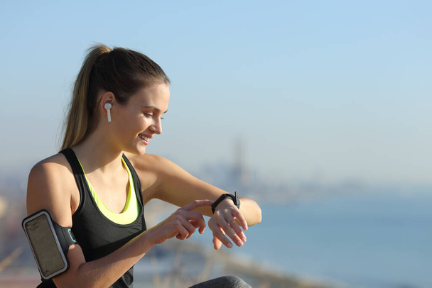 Happy runner wearing earbuds checking music on smartwatch outdoors in a city outskirts - Фото, зображення