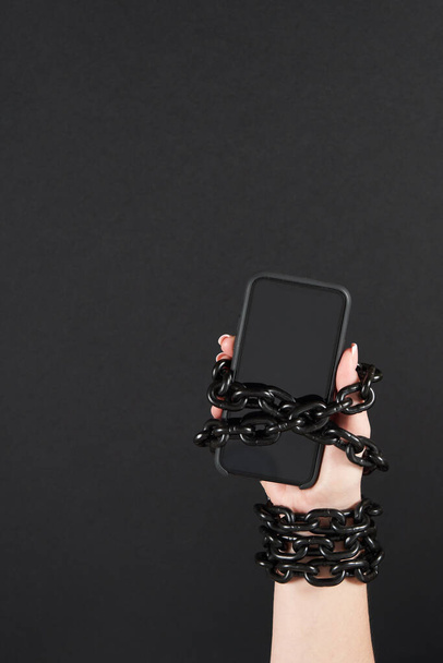 IJzeren ketting die hand en smartphone verbindt in concept van sociale media en internetverslaving een zwarte achtergrond - Foto, afbeelding