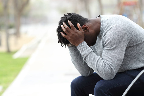 Side view portrait of a sad depressed black man sitting on a bench in a park - Foto, Imagen