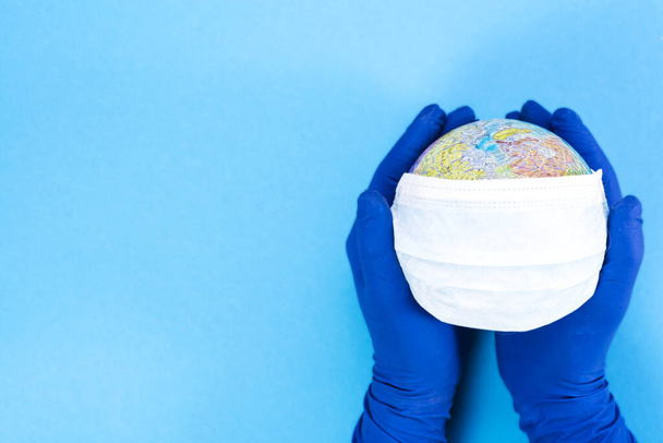 Jednorazowa maska medyczna na kuli ziemskiej w rękach lekarza na niebieskim tle, model Ziemi. Koncepcja pandemii. KOVID-19, koronawirus. Globalna epidemia choroby. COVID Niebezpieczny wirus - Zdjęcie, obraz
