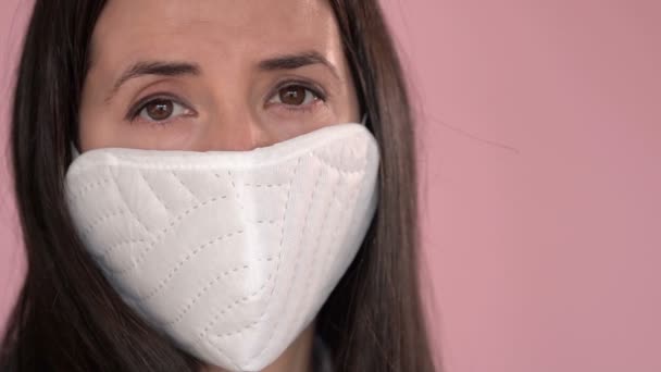 Young woman wearing a protective face mask during the Coronavirus disease (COVID-19) outbreak epidemic. Close up portrait with a multiple use white protection mask on the face shot in a studio. - Imágenes, Vídeo