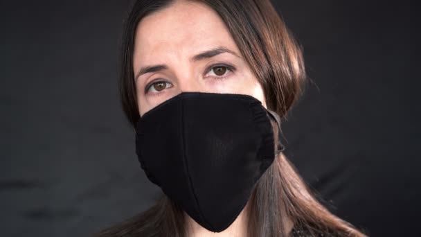 Young woman wearing a protective black leather face mask during the Coronavirus disease (COVID-19) outbreak epidemic. Close up studio portrait on a black background with a multiple use protection mask on the face. - Video