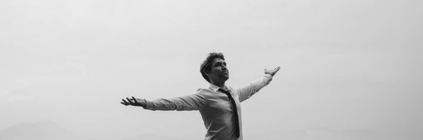 Greyscale wide view image of businessman standing under cloudy sky with his arms spread widely. - Фото, зображення