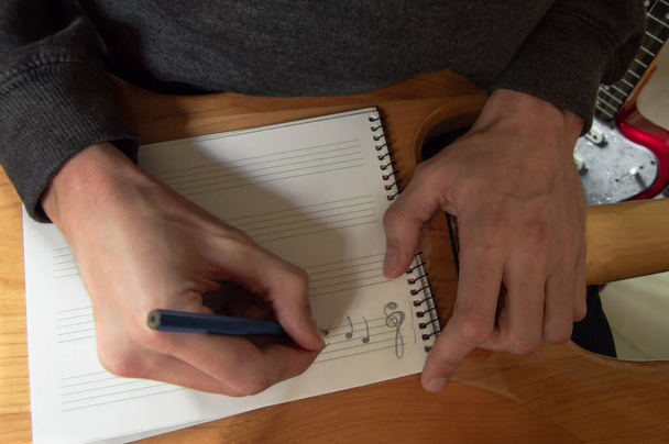 Jeune homme composant de la musique dans un cahier de notes placé sur le dos d'une guitare. Musicien écrivant dans le studio de musique. Composition en clef d'aigu
. - Photo, image