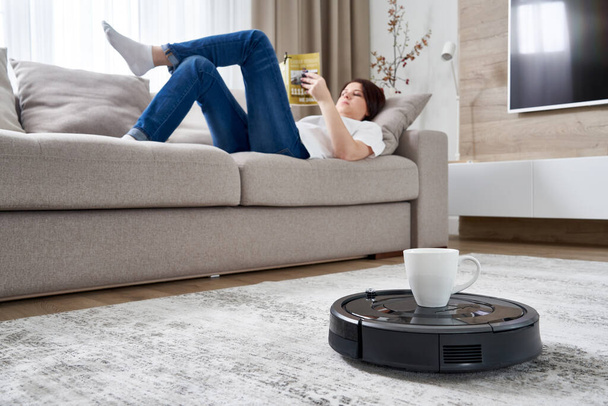 Robotic vacuum cleaner bringing cup of coffee to a woman while she is resting on sofa - Foto, Imagen