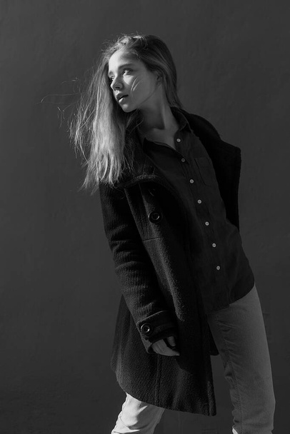Mladá krásná dívka s dlouhými vlasy v černém kabátě za slunečného dne se otočí ke zdi. Portrét stylu ulice - Fotografie, Obrázek