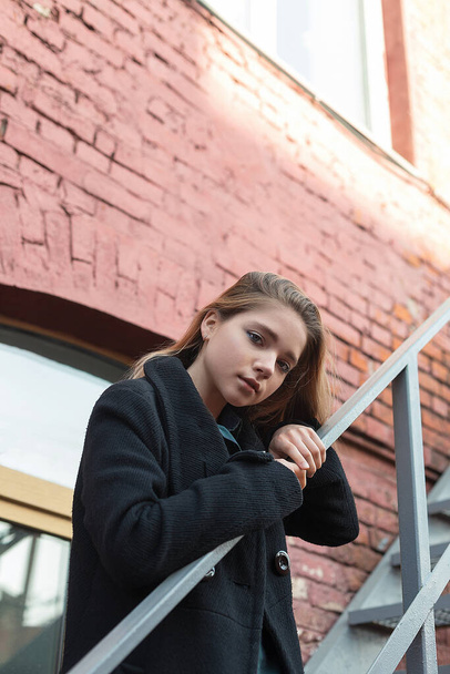 Mladá dívka v černém plášti stojí na schodech s cihlovou zdí na pozadí. Koncept osamělosti. - Fotografie, Obrázek