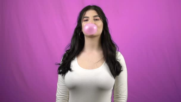 Bruneta dívka foukání velký růžový bublina žvýkačka na fialovém pozadí ve zpomaleném filmu - Záběry, video