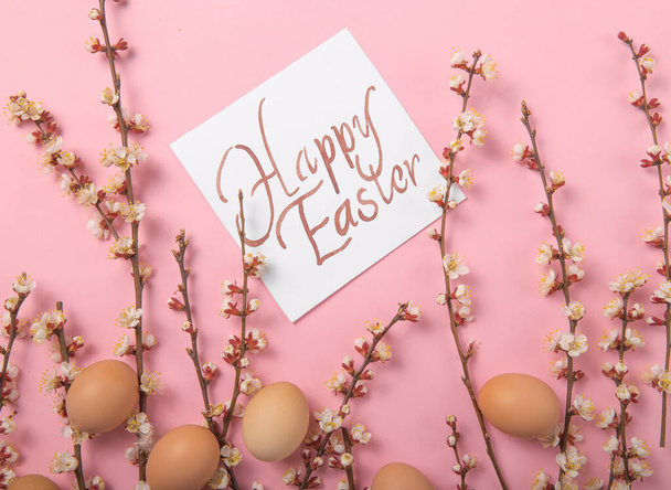Easter holiday background. Flat-lay of tender Spring  blossom flowers on branches, feathers, eggs over light pink background, top view, copy space, wide composition. Greeting card concept - Foto, imagen