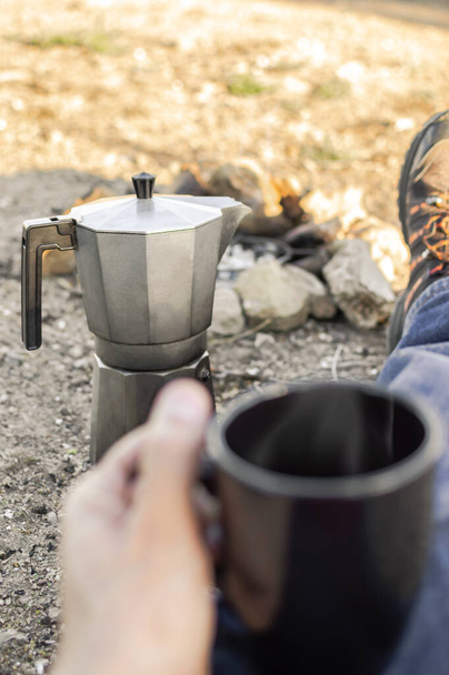 Kávékészítés tábortűzzel, pihenés a természetben.Első személytől megtekintés: tábortűz, túracipő, kávéfőző és csésze forró kávéval. - Fotó, kép