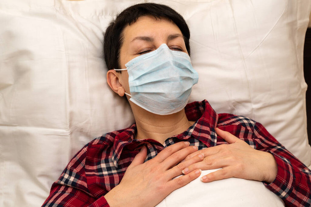 Άρρωστη νεαρή γυναίκα με ιατρική μάσκα ξαπλωμένη στο κρεβάτι να νιώθει πόνο στο στήθος και λαχάνιασμα. Μελαχρινή κοντά μαλλιά με συμπτώματα covid-19 ή πνευμονία που διαμένουν στο σπίτι κατά τη διάρκεια της πανδημίας coronavirus - Φωτογραφία, εικόνα