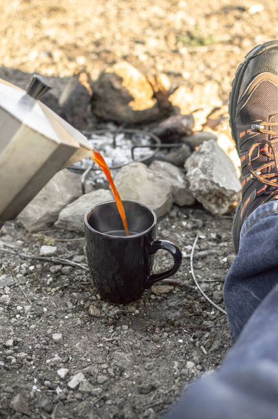 Preparo il caffe 'in falo'. L'uomo che riposa e versa caffè caldo in una tazza durante un campo nella natura. Vista in prima persona del viaggiatore seduto a terra
. - Foto, immagini