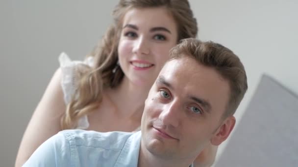 Happy beautiful couple hugging relaxing on sofa in cozy apartment looking at camera. Smiling millennial guy embracing pretty girlfriend on couch at home. Young family mortgage loan concept. Portrait - Кадры, видео
