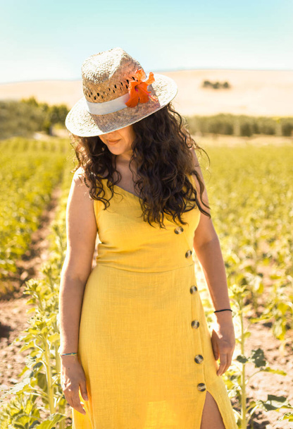 Ritratto di giovane donna dai capelli ricci neri, abito giallo e cappello di paglia con fiore in luogo naturale al tramonto. Donna guarda giù in campagna
. - Foto, immagini