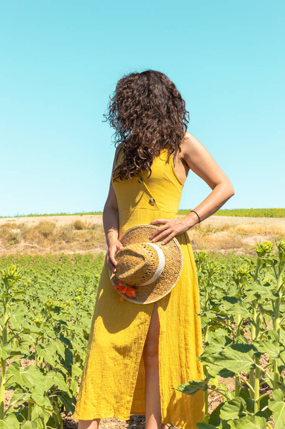 Unrecognizable woman with curly black long hair, yellow dress and straw hat in colorful natural landscape. Woman standing in rural field on sunny day. - Фото, зображення