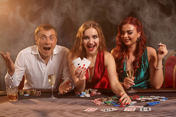 Spannende vrienden spelen poker in het casino. Ze vieren hun overwinning, glimlachen en poseren aan tafel tegen een donkere rookachtergrond. Kaarten, chips, geld, alcohol, fortuin, gokken, entertainment concept. - Foto, afbeelding