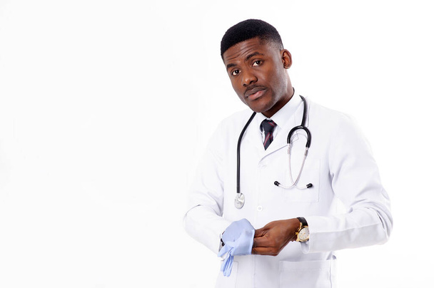Młody afrykański przystojny lekarz na białym odizolowanym tle z fonendoskopem zakłada rękawiczki medyczne - Zdjęcie, obraz