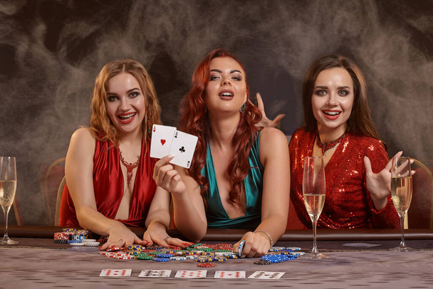 Csábító nők pókereznek a kaszinóban. Ünnepelik a győzelmüket, mosolyognak, a kamerába néznek, és sötét füstös háttérrel pózolnak az asztalnál. Kártyák, zsetonok, pénz, alkohol, szerencsejáték, szórakozás fogalma. - Fotó, kép