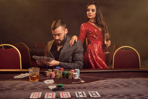 Good-looking man and gorgeous maiden are playing poker at casino. Young people are looking satisfited and counting cash winning while posing at the table against a smoke background. Cards, chips, money, gambling, entertainment concept. - Foto, imagen