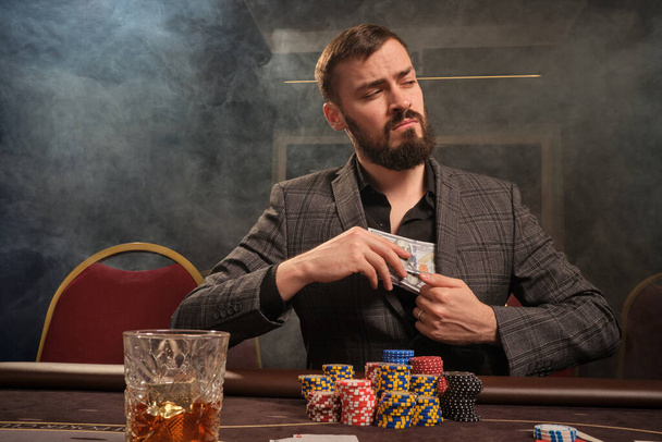 Brunet baardman in een klassiek grijs pak speelt poker in casino in rook zittend aan de tafel met chips erop. Hij verbergt wat geld in zijn jas en kijkt weg. Gokverslaving. Oprechte emoties en entertainment concept. - Foto, afbeelding