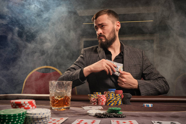 Przystojny brodaty mężczyzna w klasycznym szarym garniturze gra w pokera w kasynie z dymem, siedzi przy stole z frytkami i kartami. Ukrywa trochę gotówki w kurtce i odwraca wzrok. Uzależnienie od hazardu. Koncepcja szczerych emocji i rozrywki. - Zdjęcie, obraz