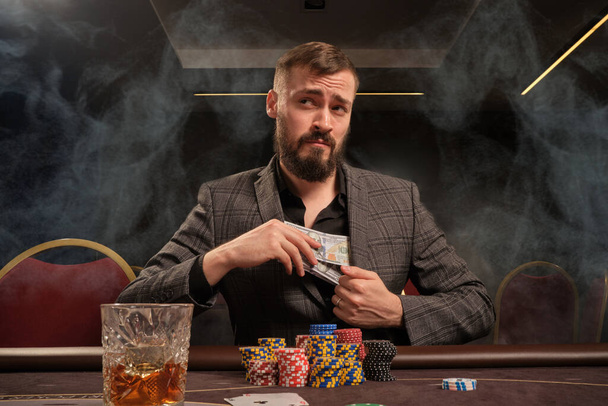 Hezký vousatý muž v klasickém šedém obleku hraje poker v kasinu v kouři, sedí u stolu s hranolkami a kartami. Schovává nějaké peníze ve své bundě a na někoho se dívá. Závislost na hazardu. Upřímné emoce a zábava con - Fotografie, Obrázek
