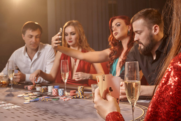 Pleasant colleagues are playing poker at casino. Youth are making bets waiting for a big win while posing at the table against a white spotlight on a dark smoke background. Cards, chips, money, alcohol, gambling, entertainment concept. - Photo, Image