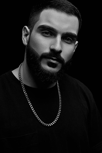 close-up of a dramatic portrait of a young serious guy, musician, singer, rapper with a beard in black clothes on a black isolated background. black and white photography. - Φωτογραφία, εικόνα