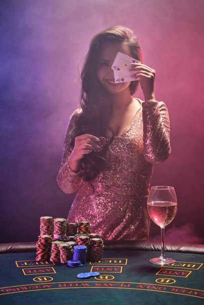 Portrait of a gorgeous brunette female with bright make-up, dressed in a golden glittered dress, posing against a gambling table with two aces and chips in her hands and closing her face with it. Poker concept on a black smoke background with pink an - Photo, Image