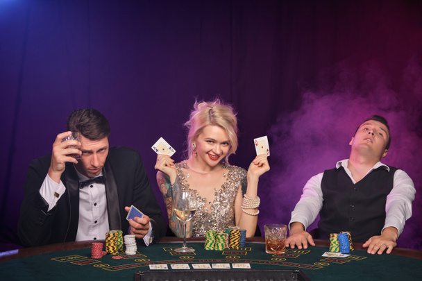 İki görkemli adam ve güzel bayan kumarhanede poker oynuyorlar. Gençler büyük bir galibiyet için bahis oynuyorlar. Siyah dumanlı arka planda kırmızı ve mavi ışıklara karşı masada oturuyorlar. Kartlar, çipler, para, kumar, eğlence c - Fotoğraf, Görsel
