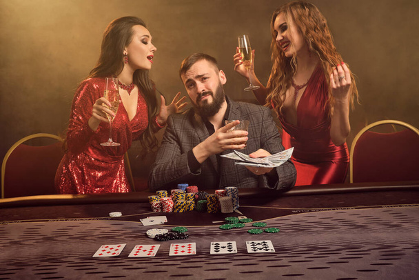 Két vonzó nő és jóképű férfi pókerezik a kaszinóban. Ők tartják a készpénz nyertes, mosolygó és jól érzik magukat, miközben pózol az asztalnál szemben a sárga háttérvilágítás füst háttér. Kártyák, zsetonok, pénz, szerencsejáték, ent - Fotó, kép