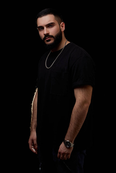 close-up of a dramatic portrait of a young serious guy, musician, singer, rapper with a beard in black clothes on a black isolated background. - Фото, изображение