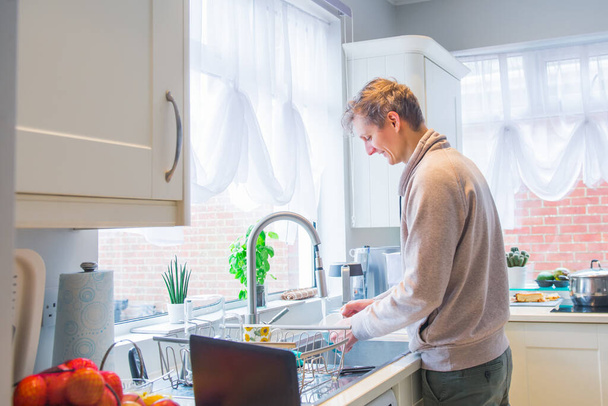 Young caucasian man working from home. Freelancer washing dishes in the kitchen and working on pc laptop. Home office, remote workplace concept. Soft selective focus. Copy space - Foto, Imagen