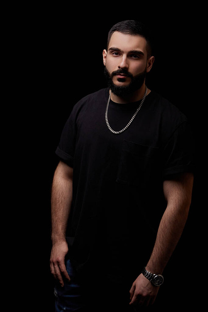 close-up of a dramatic portrait of a young serious guy, musician, singer, rapper with a beard in black clothes on a black isolated background. - Fotografie, Obrázek