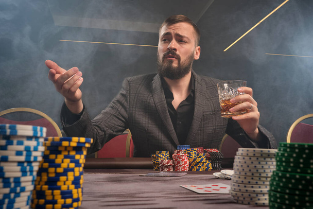 Stately bearded male in a classic gray suit is playing poker at casino in smoke sitting at the table with chips and cards on it . He is holding a glass of whiskey in his hand and looking at someone. Gambling addiction. Sincere emotions and entertainm - Photo, Image