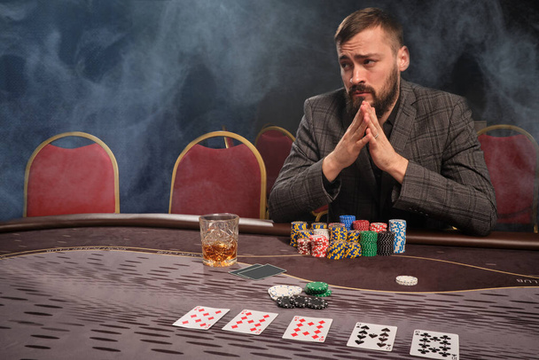 Stately bearded guy in a classic gray suit is playing poker at casino in smoke sitting at the table with chips and cards on it . Gambling addiction. Sincere emotions and entertainment concept. - Photo, Image
