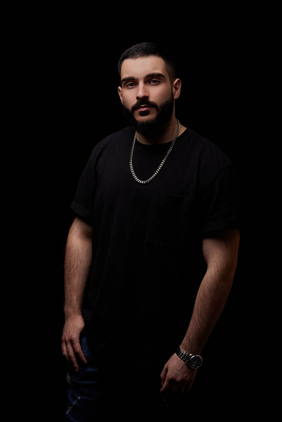 close-up of a dramatic portrait of a young serious guy, musician, singer, rapper with a beard in black clothes on a black isolated background. - Foto, Bild