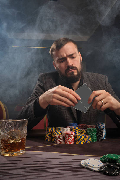 Brunet brodaty mężczyzna w klasycznej szarej marynarce gra w pokera w kasynie z dymem, siedzi przy stole z frytkami i szklanką whisky na nim. Patrzy na karty w ręku. Uzależnienie od hazardu. Koncepcja rozrywki. - Zdjęcie, obraz