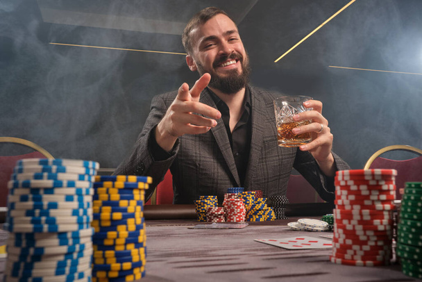 Athletic bearded person in a classic gray suit is playing poker at casino in smoke sitting at the table with chips and cards on it . He is holding a glass of whiskey in his hand and pointing at you. Gambling addiction. Sincere emotions and entertainm - Photo, Image