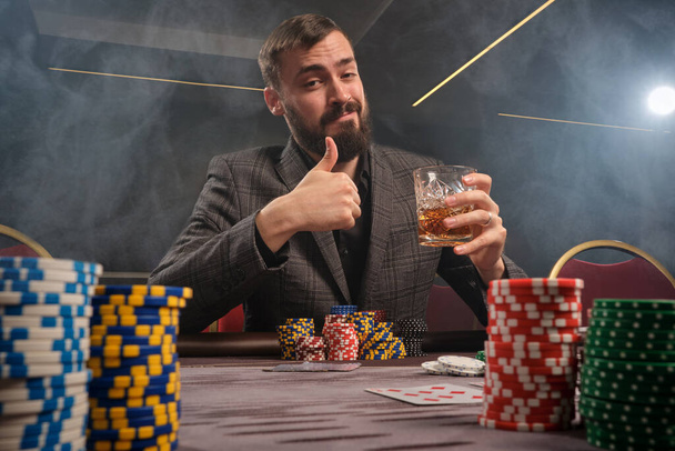 Athletic bearded fellow in a classic gray suit is playing poker at casino in smoke sitting at the table with chips and cards on it . He is holding a glass of whiskey in his hand and showing thumb up while looking at the camera. Gambling addiction. Si - Photo, Image