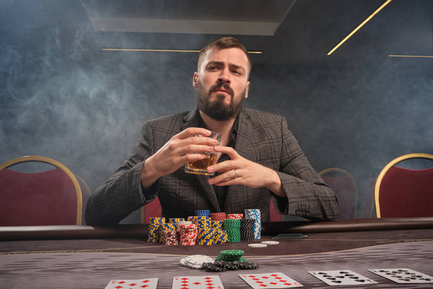 Stylish bearded fellow in a classic gray suit is playing poker at casino in smoke sitting at the table with chips and cards on it . He is holding a glass of whiskey in his hand and looking at the camera. Gambling addiction. Sincere emotions and enter - Photo, Image