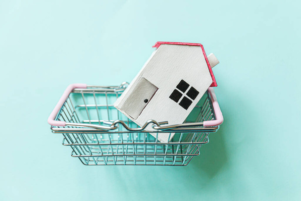 Simply design with miniature white toy house and supermarket grocery shopping basket isolated on blue background. Mortgage property insurance dream home concept. Copy space - Photo, Image