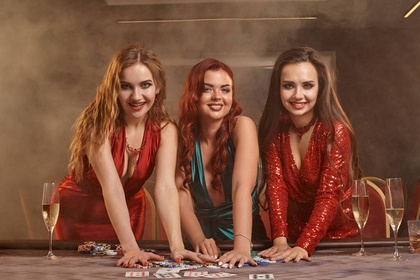 Three beautiful women are playing poker at casino. They are celebrating their win, looking at the camera and smiling, holding some chips and posing at the table against a smoke background. Cards, money, alcohol, gambling, entertainment concept. - Foto, imagen